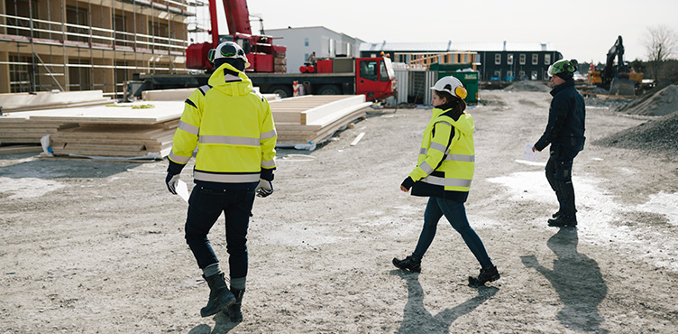 Tre personer syns vid byggarbetsplats, två av dom är klädda i neonfärgade jackor och hjälm.
